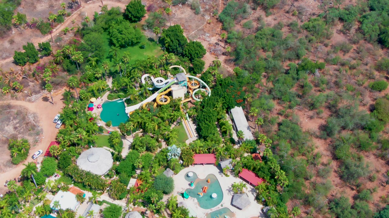 Aerial frame in Caduano showing the water slide tower and curving tubes above a green pool area, with dry vegetation across the background and small vehicles along the dirt access road.