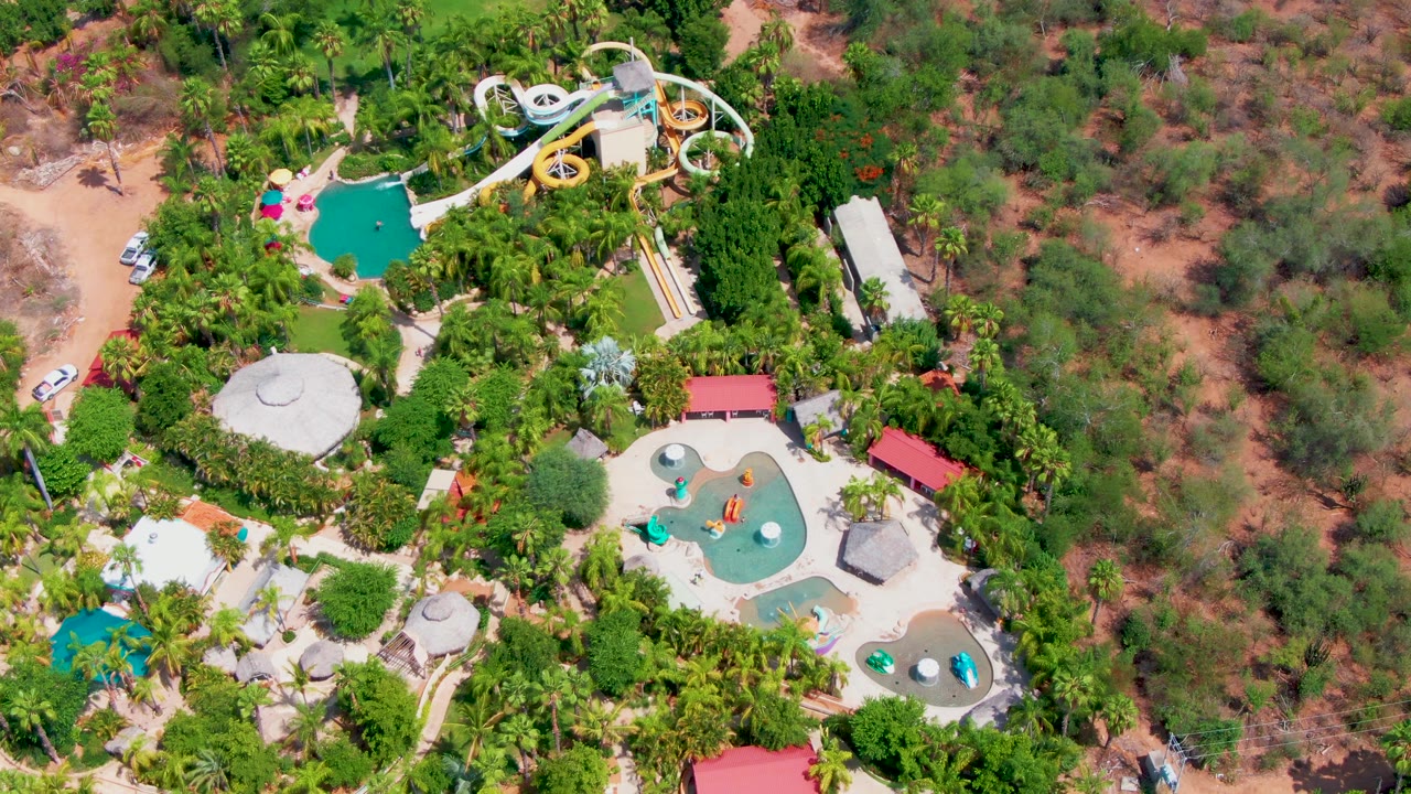 Overhead view of a Caduano, Baja California Sur water park with turquoise pools, looping slides, dense palms, sandy access areas, a small car near the edge, and no visible people.