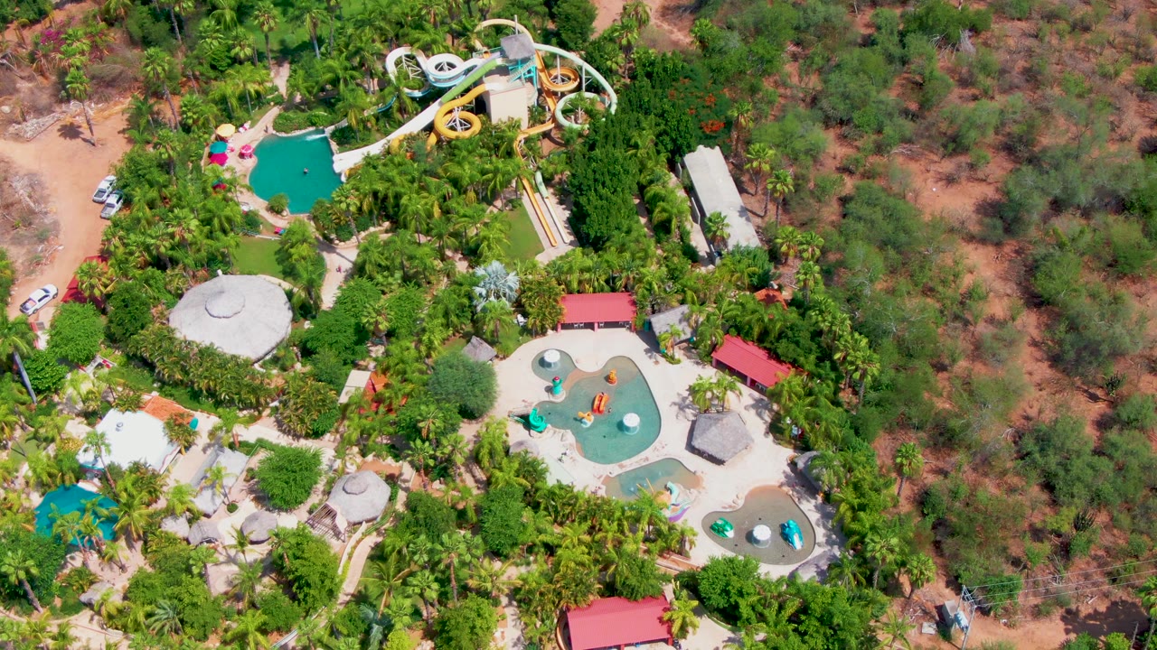 Aerial frame over Caduano showing the slide complex and pools set inside thick greenery, with scrubby brown terrain to the right and no visible people.