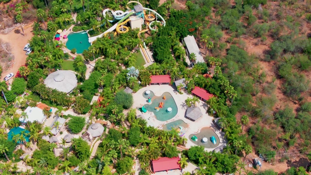 High drone view in Caduano, Baja California Sur with curving water slides at the top, several pools below, red-roofed buildings, sandy terrain, and no visible people.