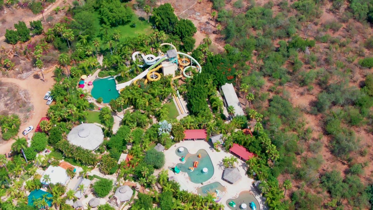 Aerial frame over Caduano showing the water slide near the top of the park, pools below it, thick green landscaping, scrubby terrain, and a small road edge, with no visible people.