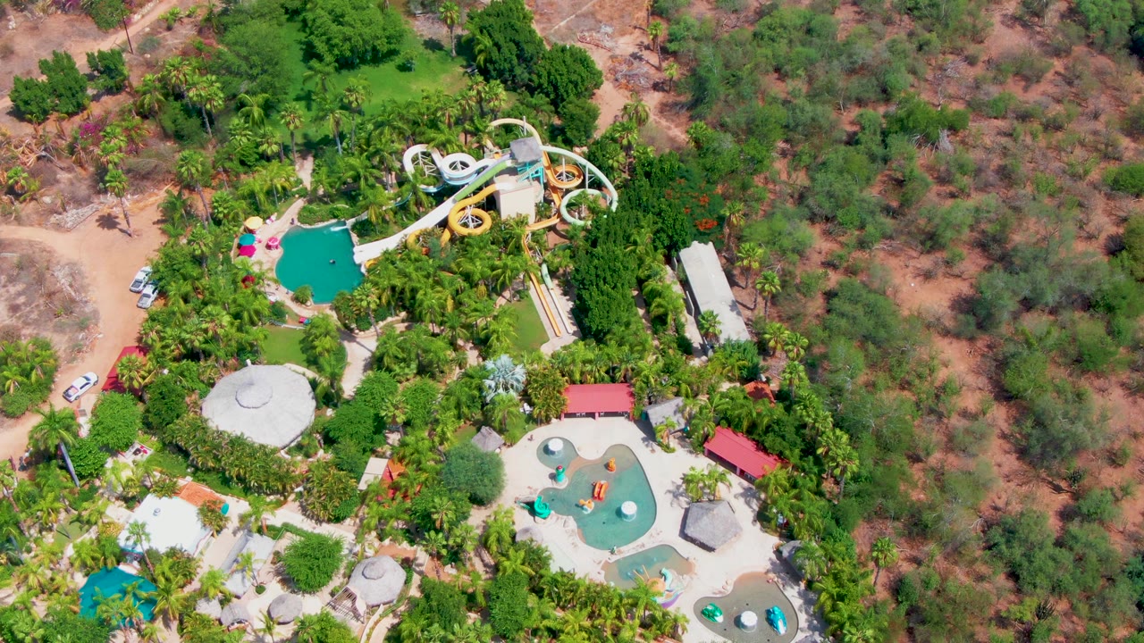 Overhead Caduano water park view with the slide complex high in frame, turquoise pool basins lower in frame, palms and trees around the grounds, and dry vegetation beyond, with no visible people.