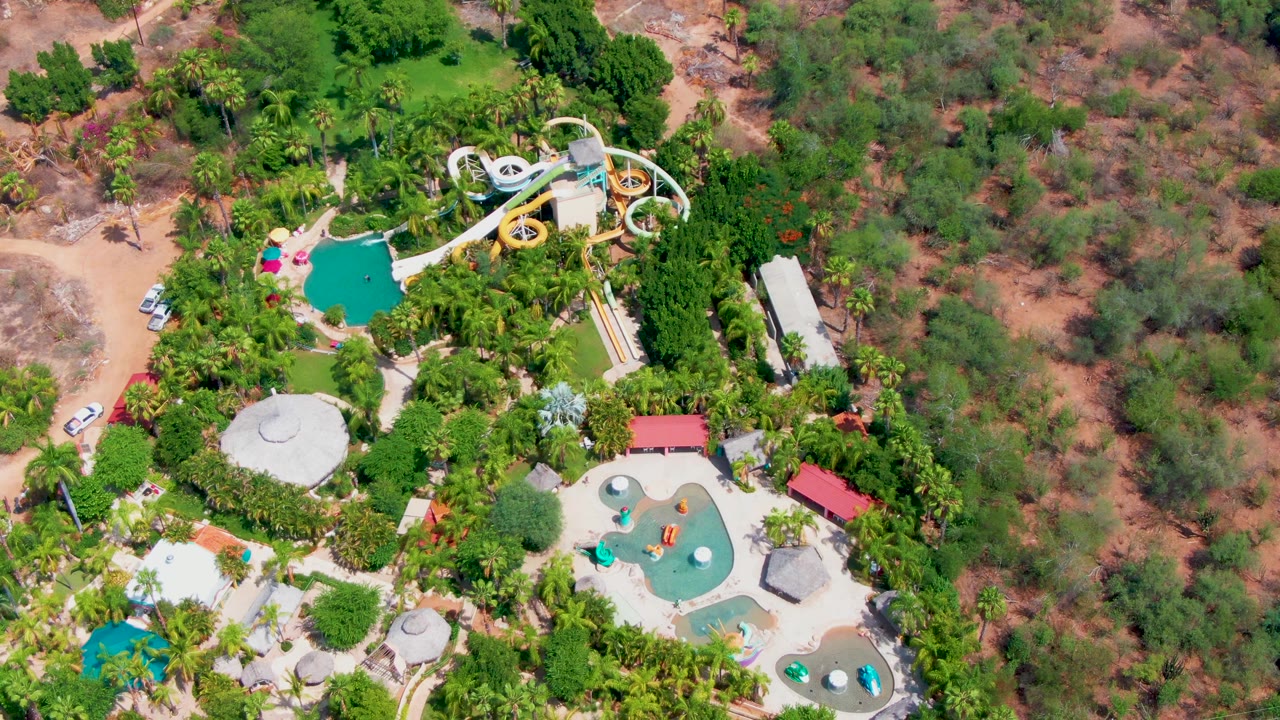 Drone-style overhead frame in Caduano with the yellow-and-white slide and upper pool near the top, lower pools and shaded structures below, a road at left, and no visible people.