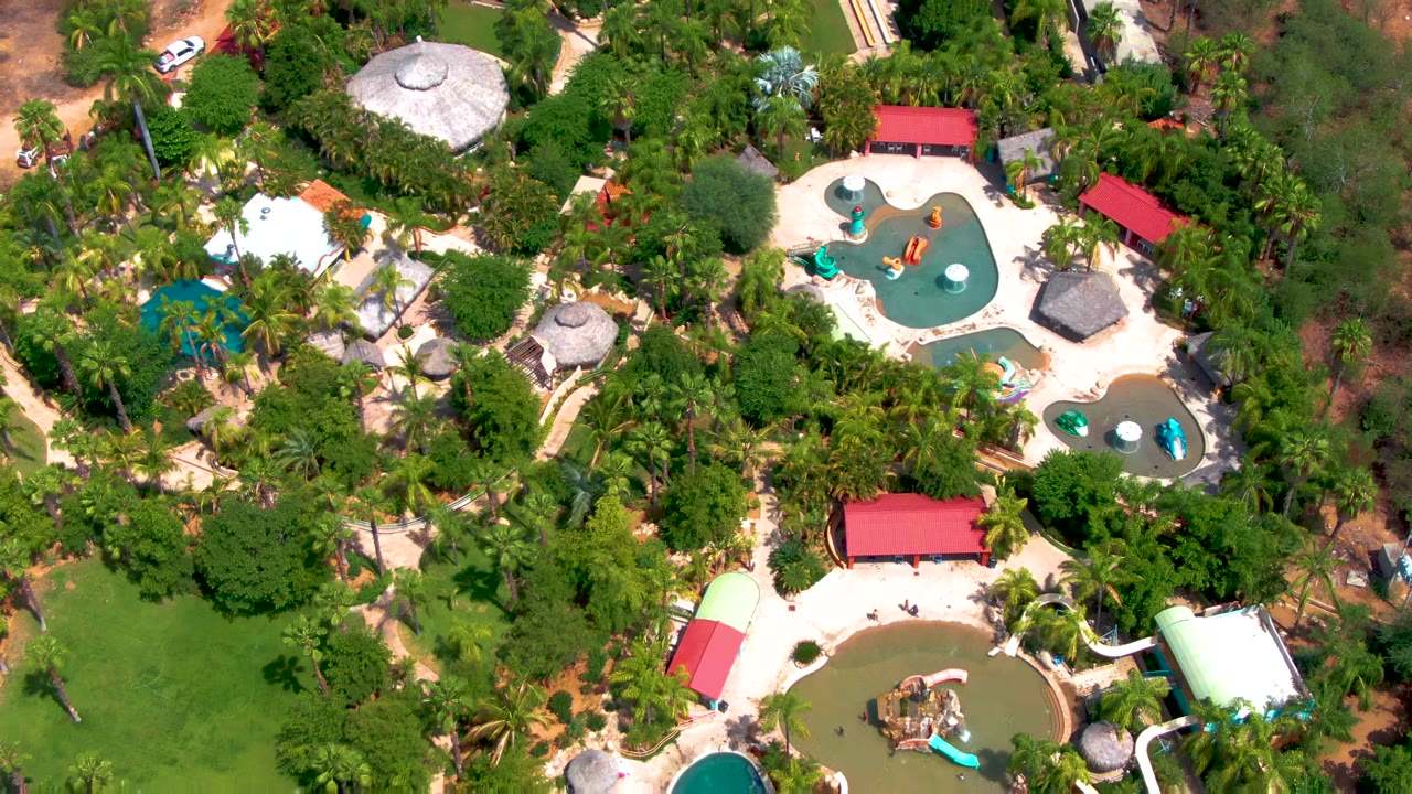 Overhead view of a tropical water park in Caduano, Baja California Sur, with turquoise pools, curved slides, red roofs, palms, and small people near the pool decks.