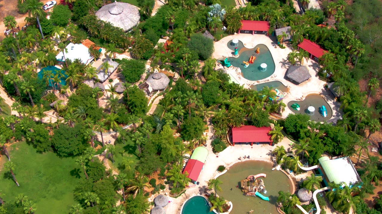 The frame presents the Caduano water park from overhead, with red-roofed shelters, rounded pools, slide structures, thick green planting, and visitors reduced to tiny figures.