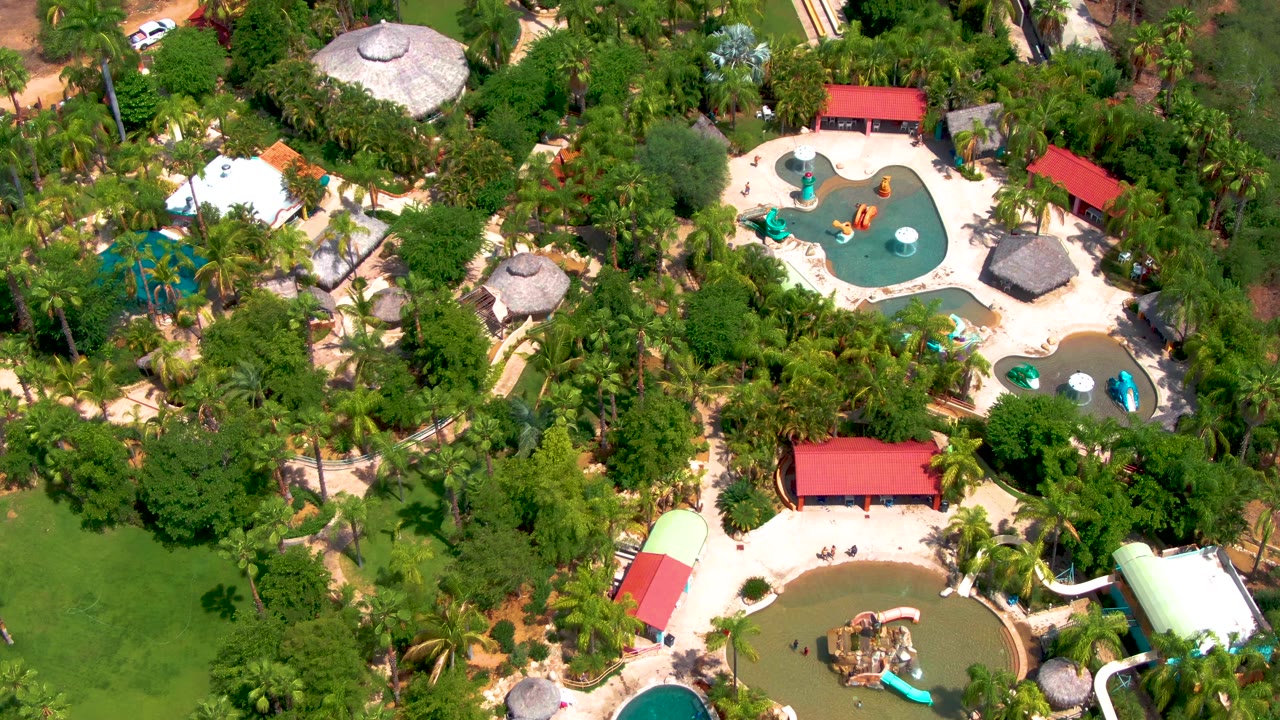 High aerial frame over a Caduano water park in Baja California Sur, Mexico, showing turquoise pools, orange and blue slide sections, red roofs, dense palms, and no visible people.
