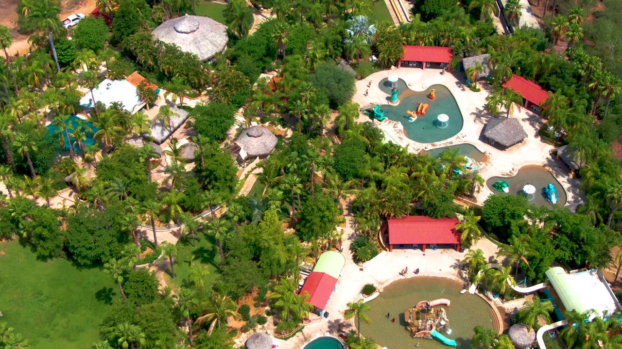 Second overhead frame of the Caduano water park, with the curving turquoise pool near the upper right, red roofs across the middle, slide structures below, and no visible people.