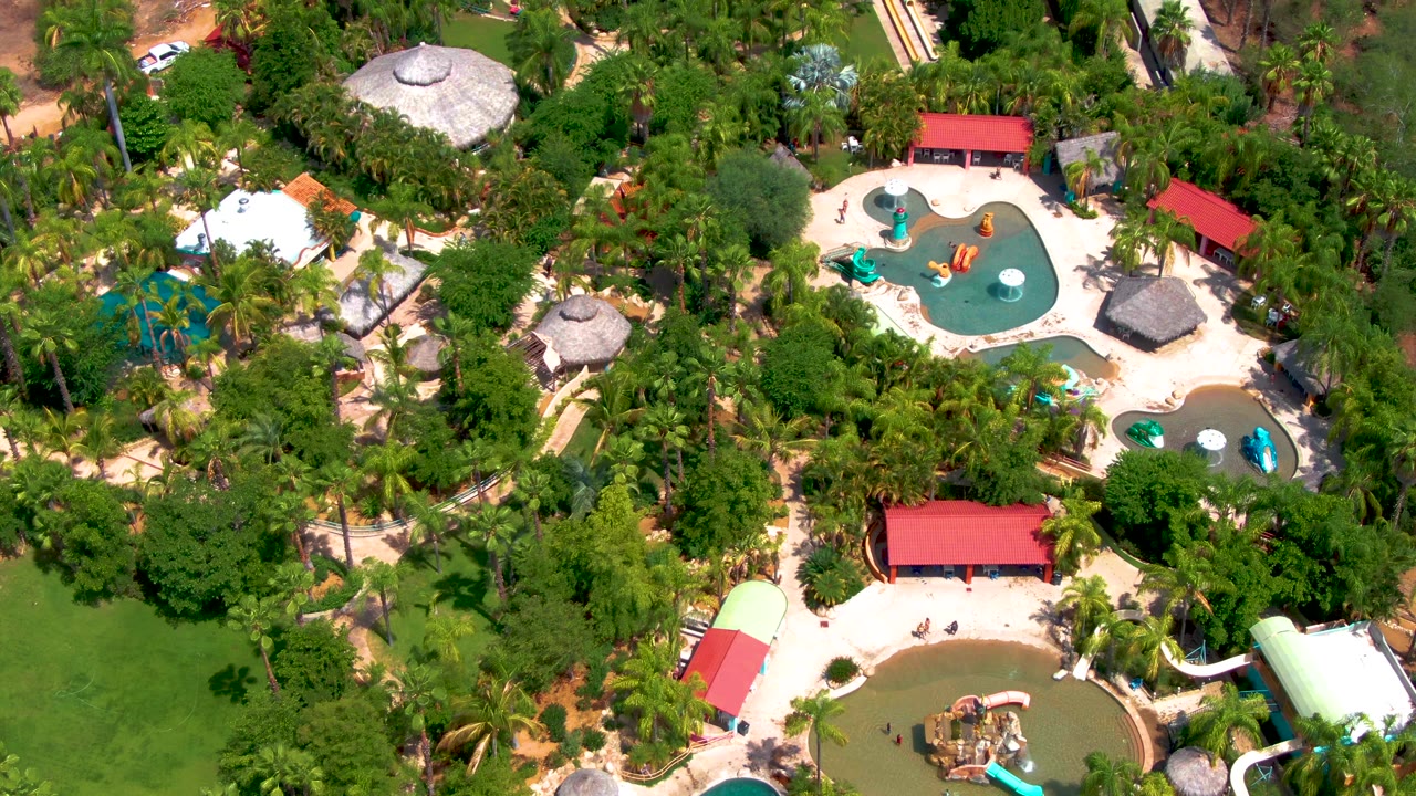 Later overhead frame in Caduano showing the water park partly screened by palms, with a turquoise pool at upper right, a brown pool at lower edge, red rooflines, and no visible people.