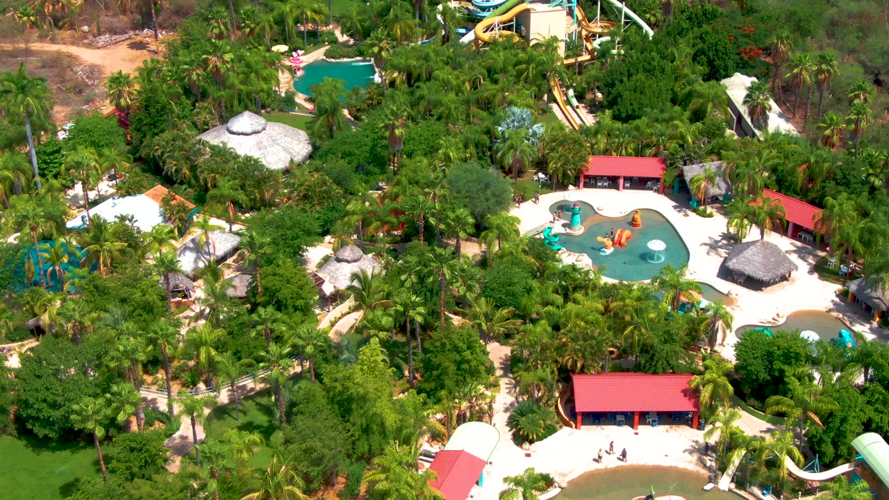 Overhead view of a water park in Caduano, Baja California Sur, with turquoise pools, yellow and white slides, red-roofed buildings, thatched shade roofs, dense palms, and no visible people.
