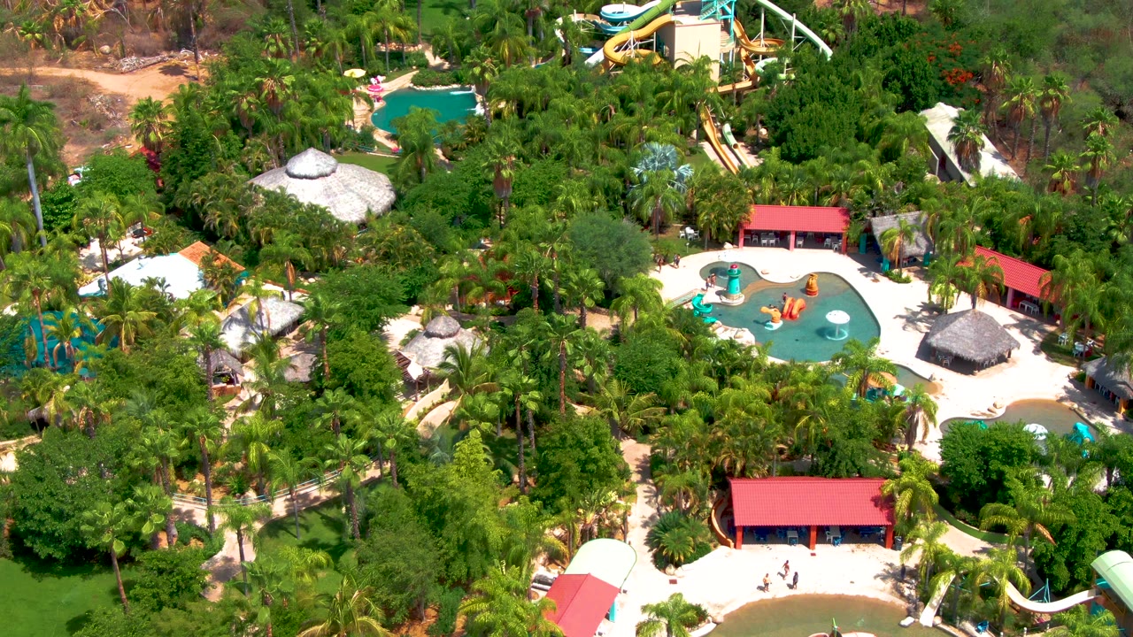Aerial frame over Caduano's water park showing the central blue pool, curved slide features, red roofs, pale deck areas, tree canopy, and no visible people.