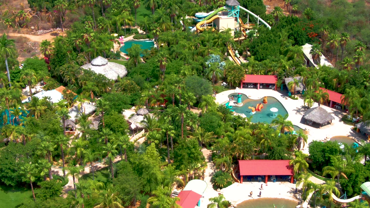 Aerial water park scene with pools, thatched roofs, palms, and red buildings.
