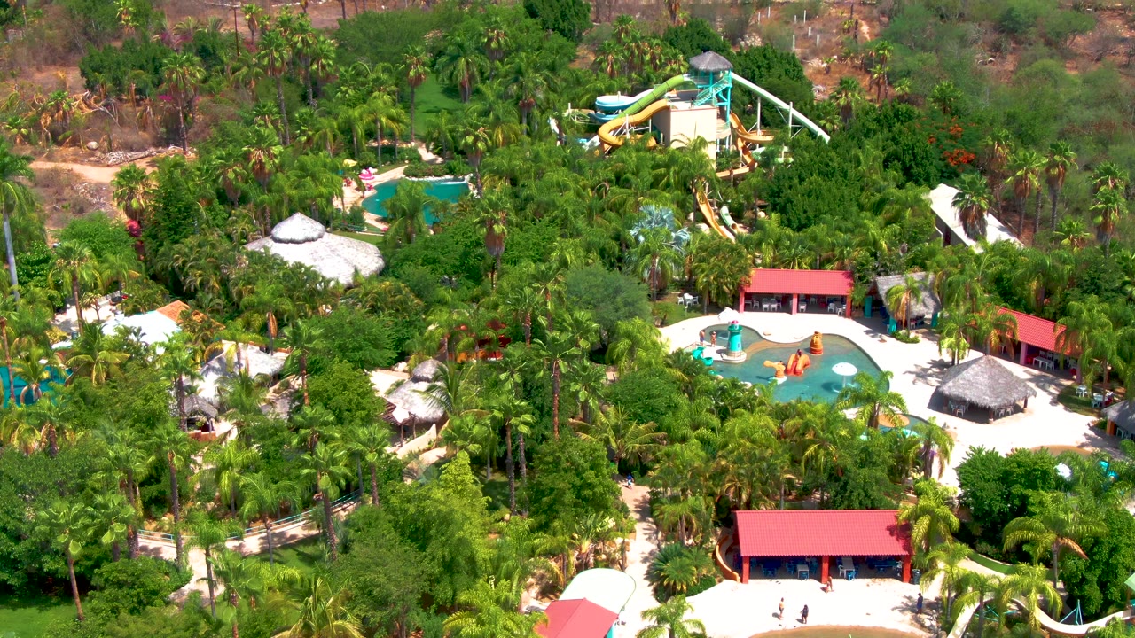Aerial view over a Caduano, Baja California Sur water park with blue pools, yellow slides, red roofs, palms, and dry hillside beyond, with no visible people.