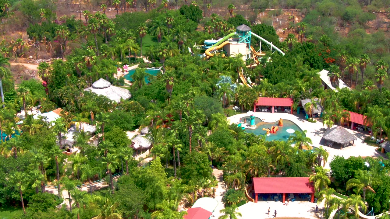 Drone view in Caduano showing the slide tower near the upper frame, blue pool area below it, red-roofed structures, and thick palm cover, with no visible people.