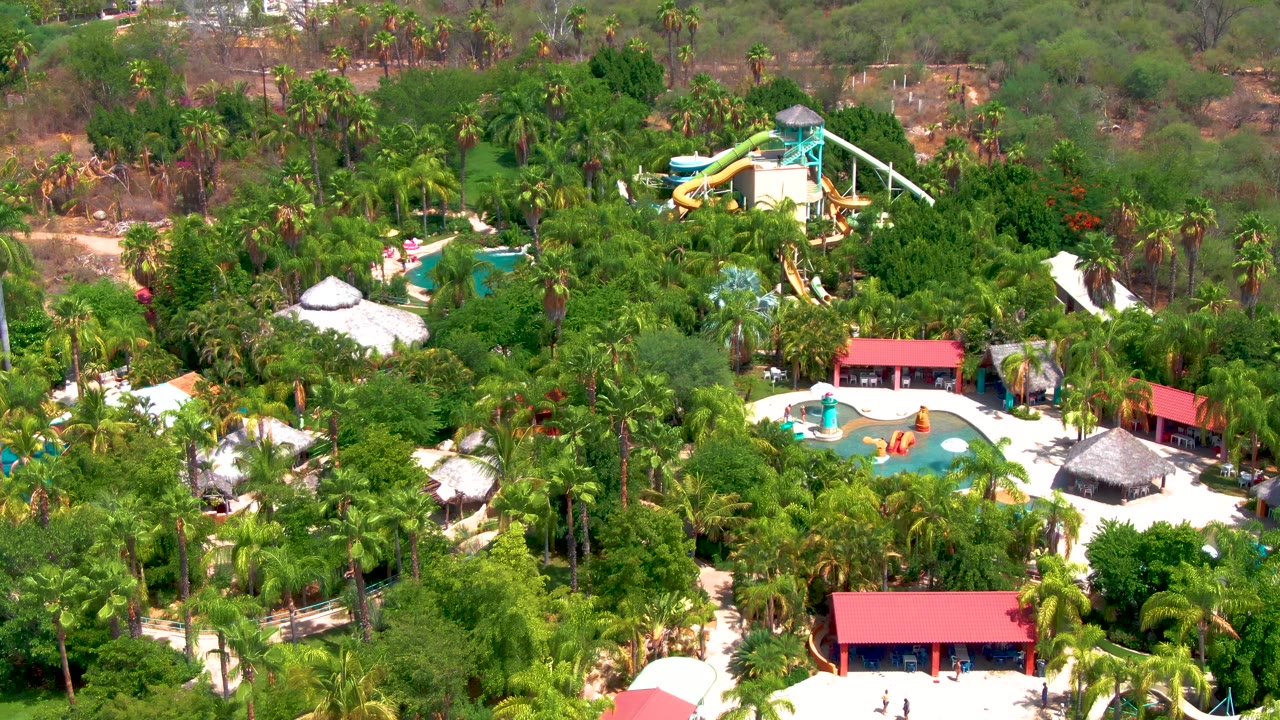 High aerial frame of the Caduano water park with curved blue pools, yellow slide features, red roofs along the lower area, palms, and scrubby hillside behind, with no visible people.