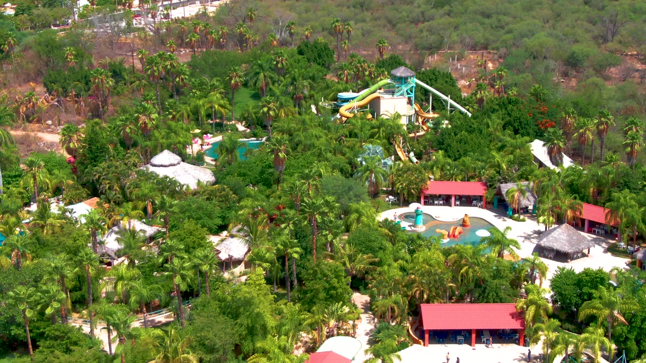Aerial composition over Caduano with the water slide tower set against dense green tree cover, pools and red-roofed buildings lower in frame, and no visible people.