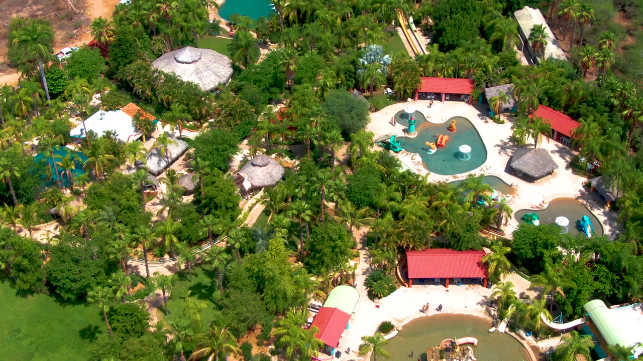 Overhead view of a Caduano, Baja California Sur water park with dense trees, turquoise pools, thatched roofs, red-roofed buildings, and small visible people near the pools.