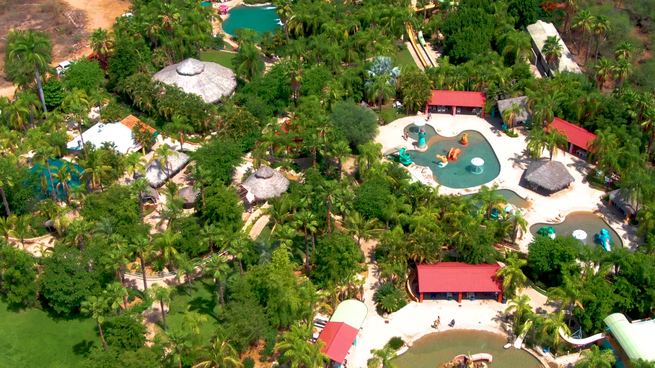 The Caduano, Baja California Sur water park remains framed from above, with blue-green pools, a curved yellow slide, red roofs, white paths, and palms filling most of the view.