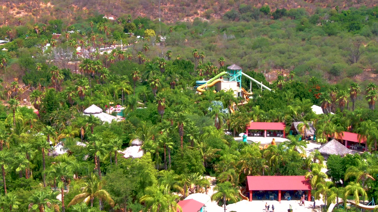 Wide aerial frame over a Caduano, Baja California Sur resort with a yellow water slide tower rising above dense palms, red roofs below, dry hills behind, and no clearly visible people.