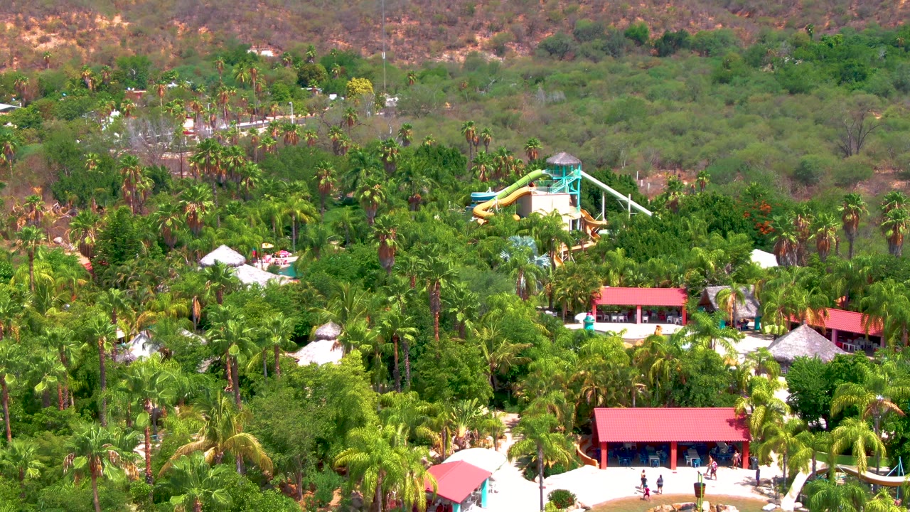 Elevated resort view with palms, red roofs, water slide tower, and hillside background.