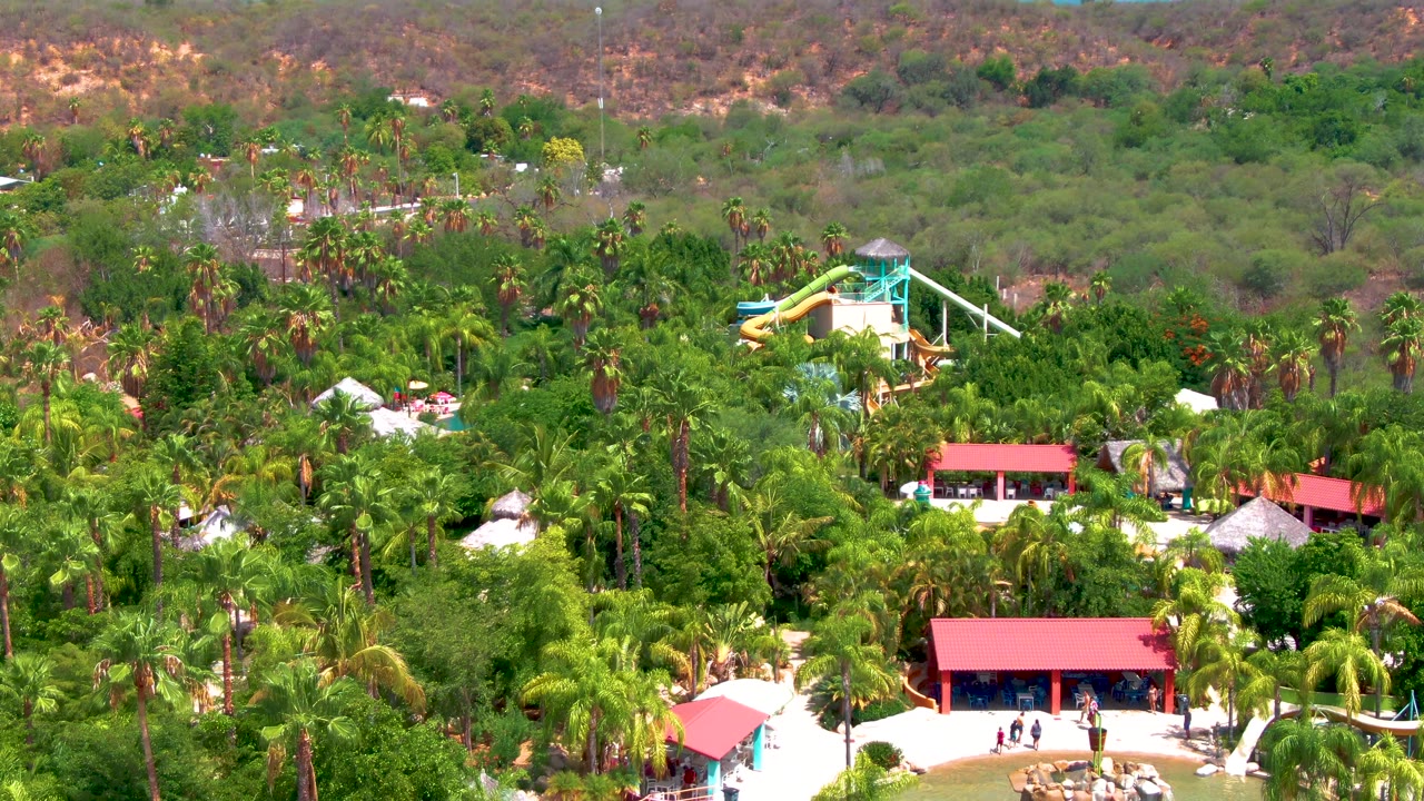 High wide view of the Caduano resort landscape, with the slide tower slightly right of center, red roofs at the lower edge, a thick palm canopy, and dry hills in the distance.