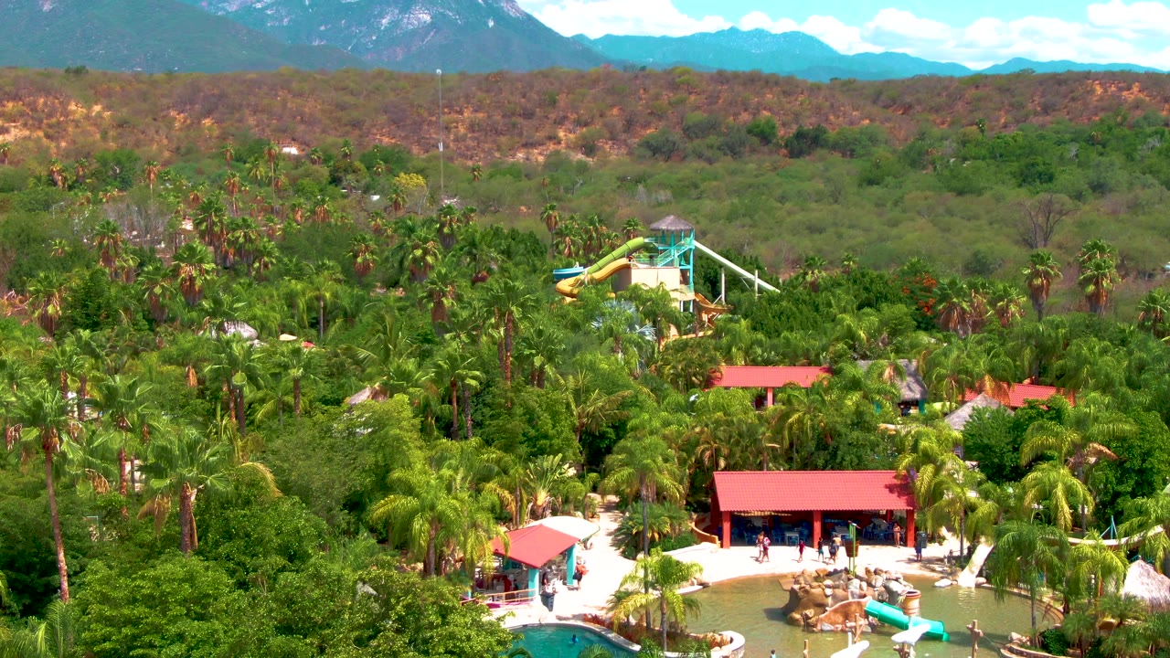 Aerial frame over a Caduano water park with red-roofed pool buildings, palms, green and yellow slides, distant hills, mountains, blue sky, and small people visible near the pool area.