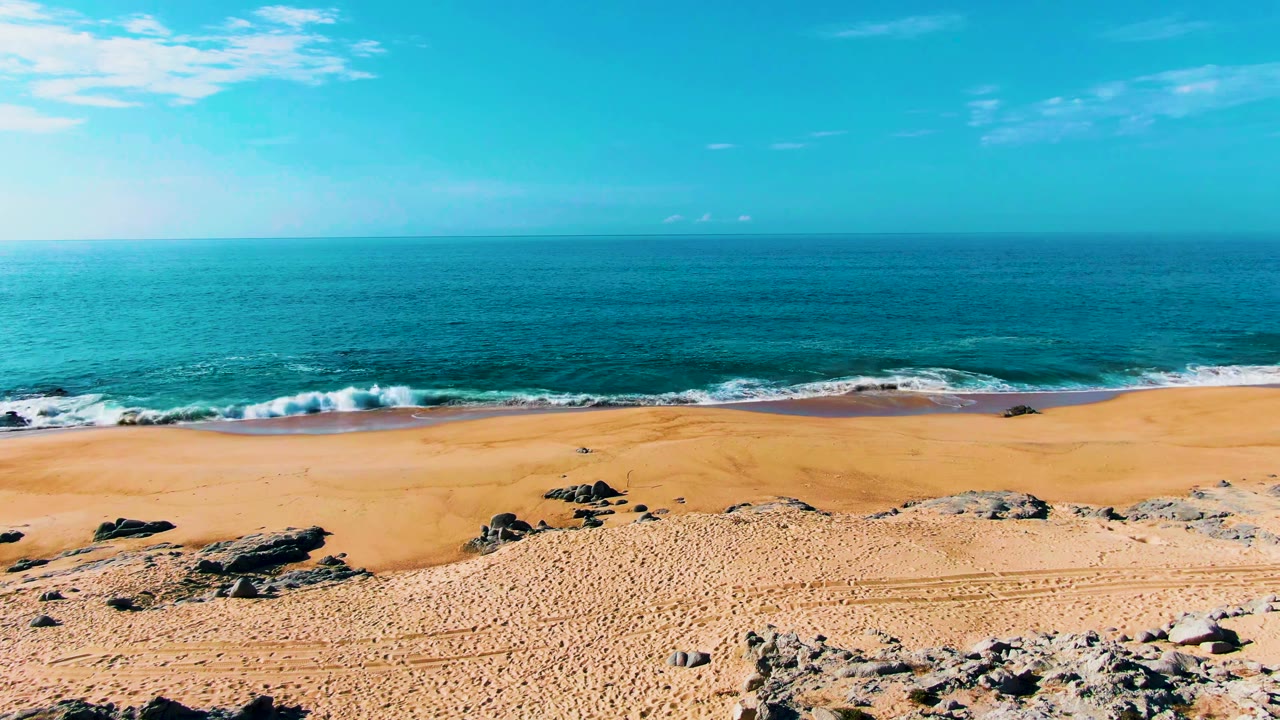 Opening view of Buzzards Beach in Baja California Sur, Mexico, with sandy foreground rocks, small breaking waves, a blue ocean horizon, and no visible people.