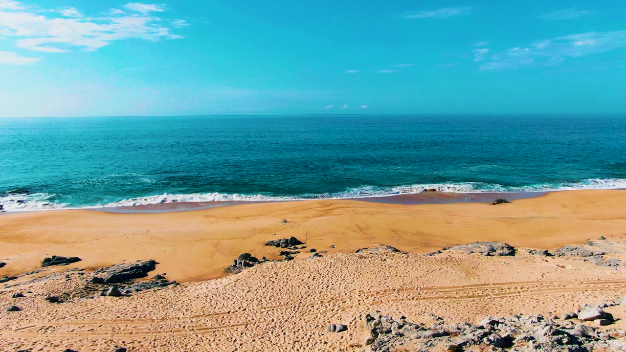 Early frame of the Punta Buzzards shoreline with rock clusters across the sand, turquoise water rolling in, thin clouds above, and no visible people.