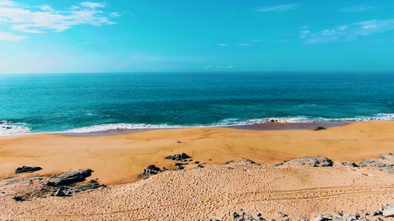 The beach view continues at Buzzards Beach, with scattered dark rocks, textured sand, a narrow band of white foam, deep blue water, and no visible people.