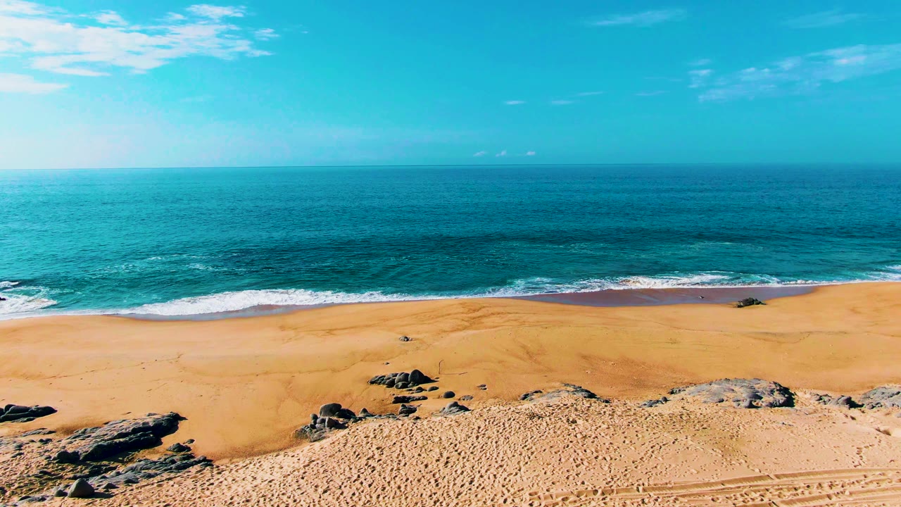 Mid-clip view of Punta Buzzards with open golden sand, fewer rocks in the center, gentle surf curling along the shoreline, and no visible people.