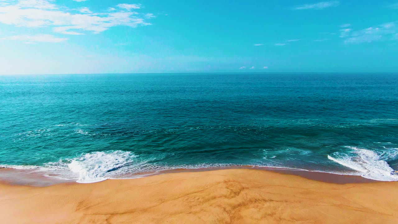 Aerial view at Punta Buzzards, Baja California Sur, with turquoise ocean meeting a sandy shoreline, white foam near the lower edge, and no visible people.
