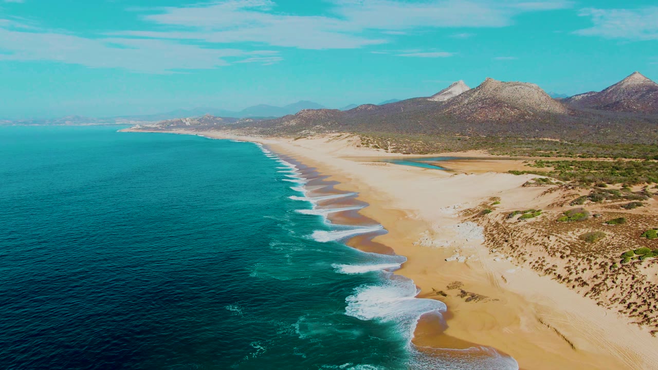Aerial view of Buzzards Beach in Baja California Sur with turquoise water on the left, waves breaking onto sand, dunes inland, mountains beyond, and no visible people.
