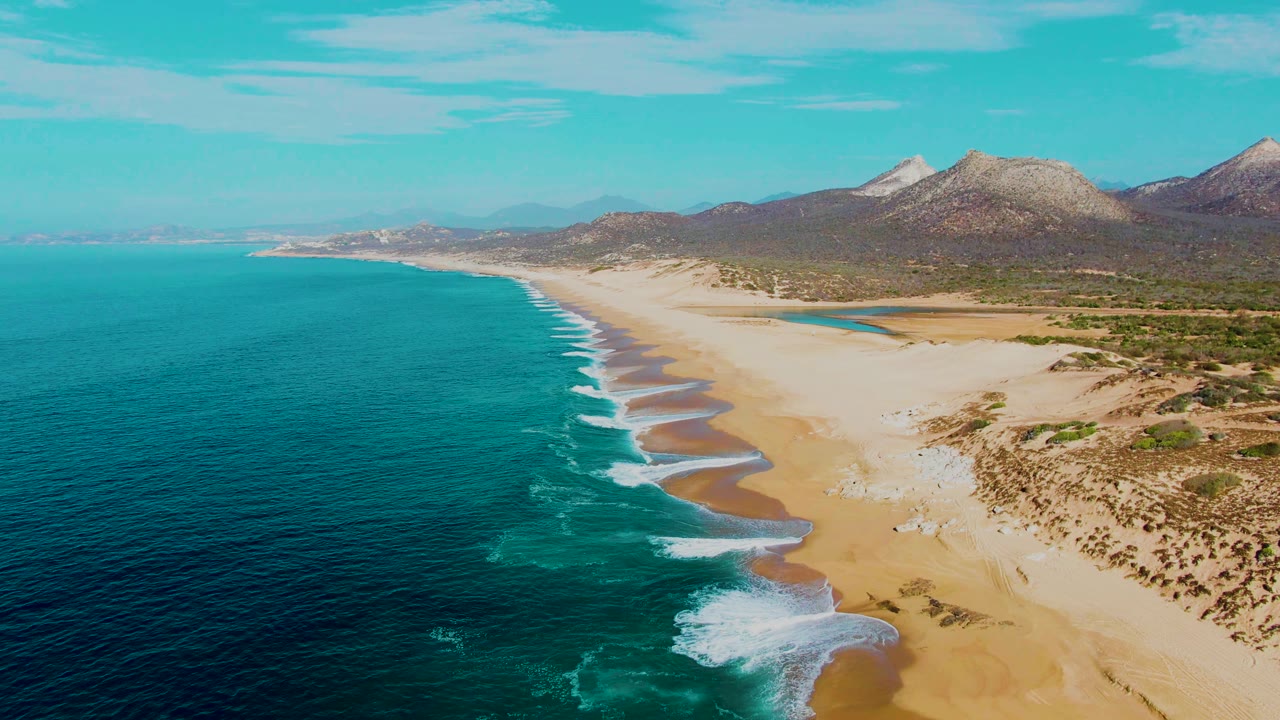 Wide aerial beach view with surf bands and mountains in the distance.