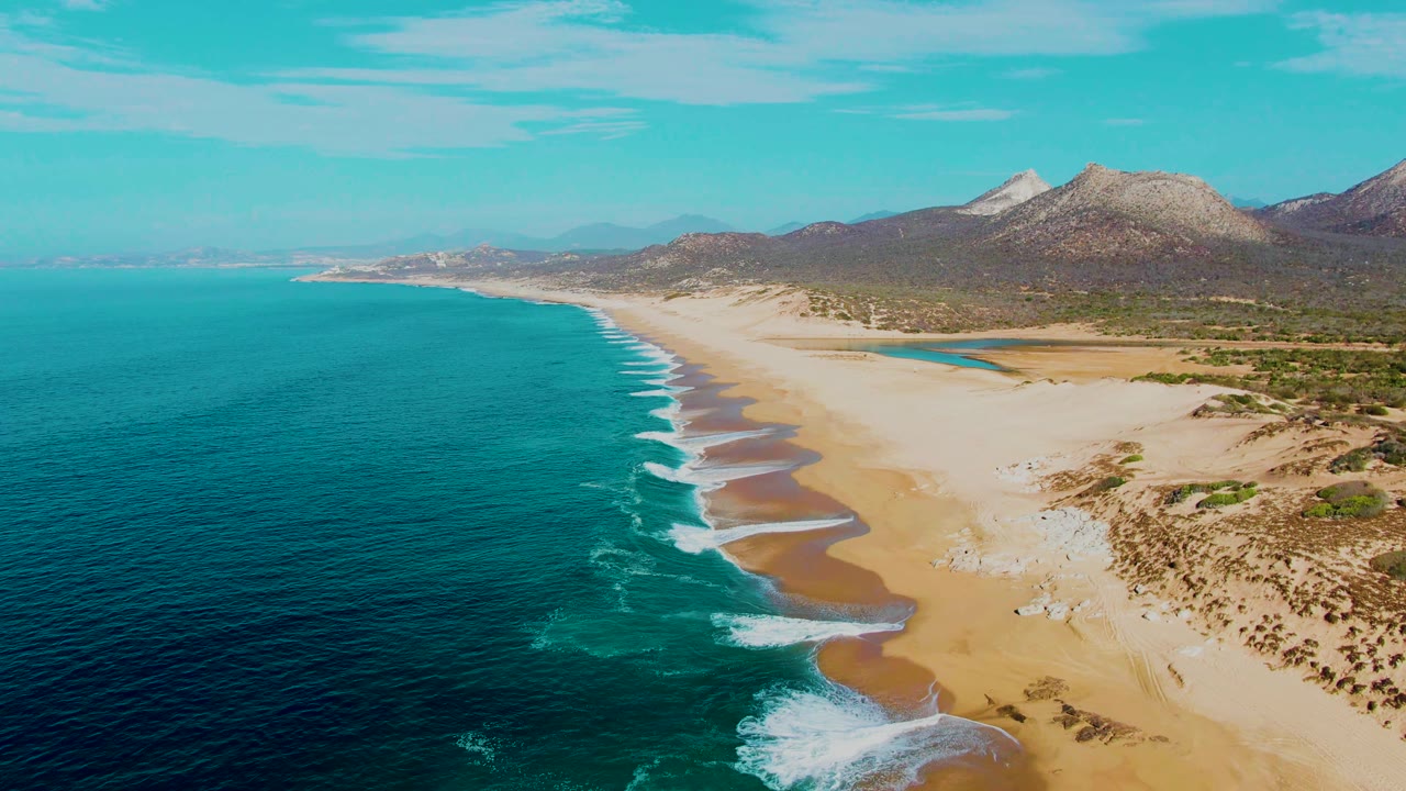 Aerial frame over Buzzards Beach with curved surf lines reaching the beach, tan sand and dune textures to the right, arid mountains on the horizon, and no visible people.