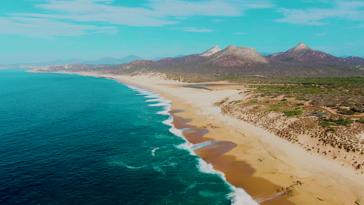Opening aerial view of Punta Buzzards coastline in Baja California Sur, with deep blue ocean on the left, white surf on sand, dunes and mountains beyond, and no visible people.
