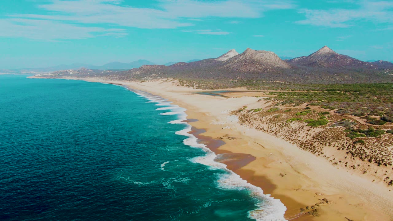 Punta Buzzards aerial frame showing white wave bands advancing toward the sandy shore, with turquoise shallows, darker ocean water, low vegetation, mountains, and no visible people.