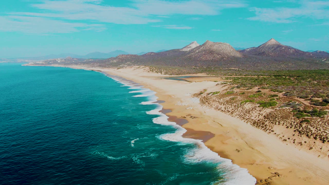 Early clip frame over Punta Buzzards with the shoreline running from lower right to upper left, surf foam reaching the sand, desert scrub inland, mountains on the horizon, and no visible people.