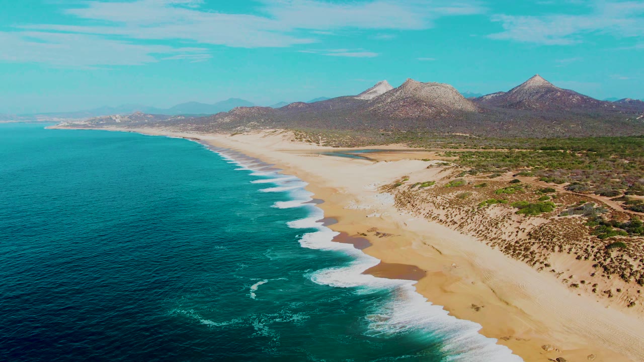 Drone view of sandy coast, rolling surf, scrubland, and distant rocky mountains.