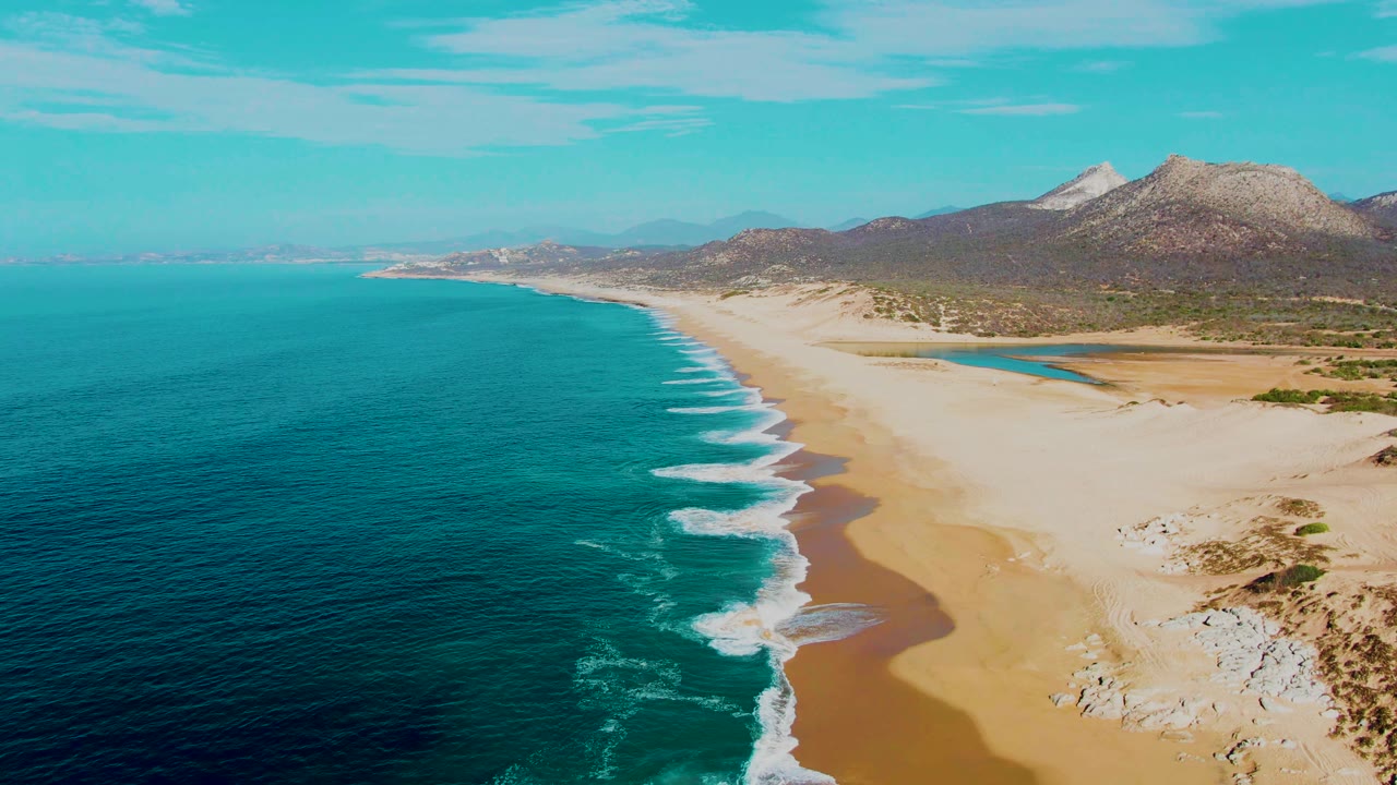 Opening aerial frame over Buzzards Beach in Baja California Sur, showing turquoise water on the left, a pale sandy shoreline, distant mountains, clear sky, and no visible people.