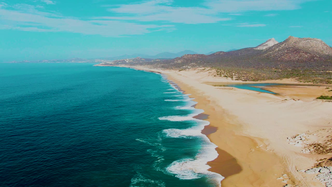 Aerial frame from Punta Buzzards showing white surf curling into shallow water, a wide tan beach, rocky sand patches near the lower right, mountains, and no visible people.