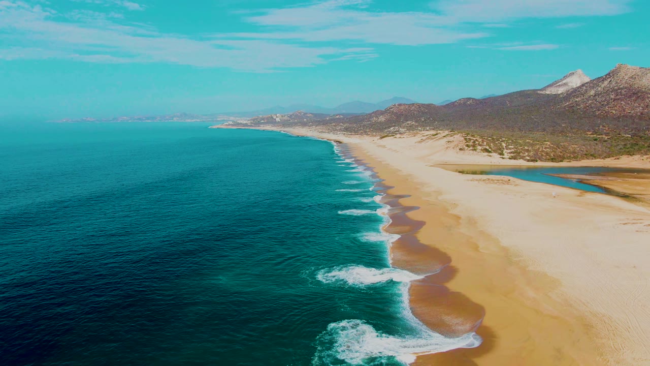 Aerial view of turquoise ocean waves beside a wide sandy beach and mountains.