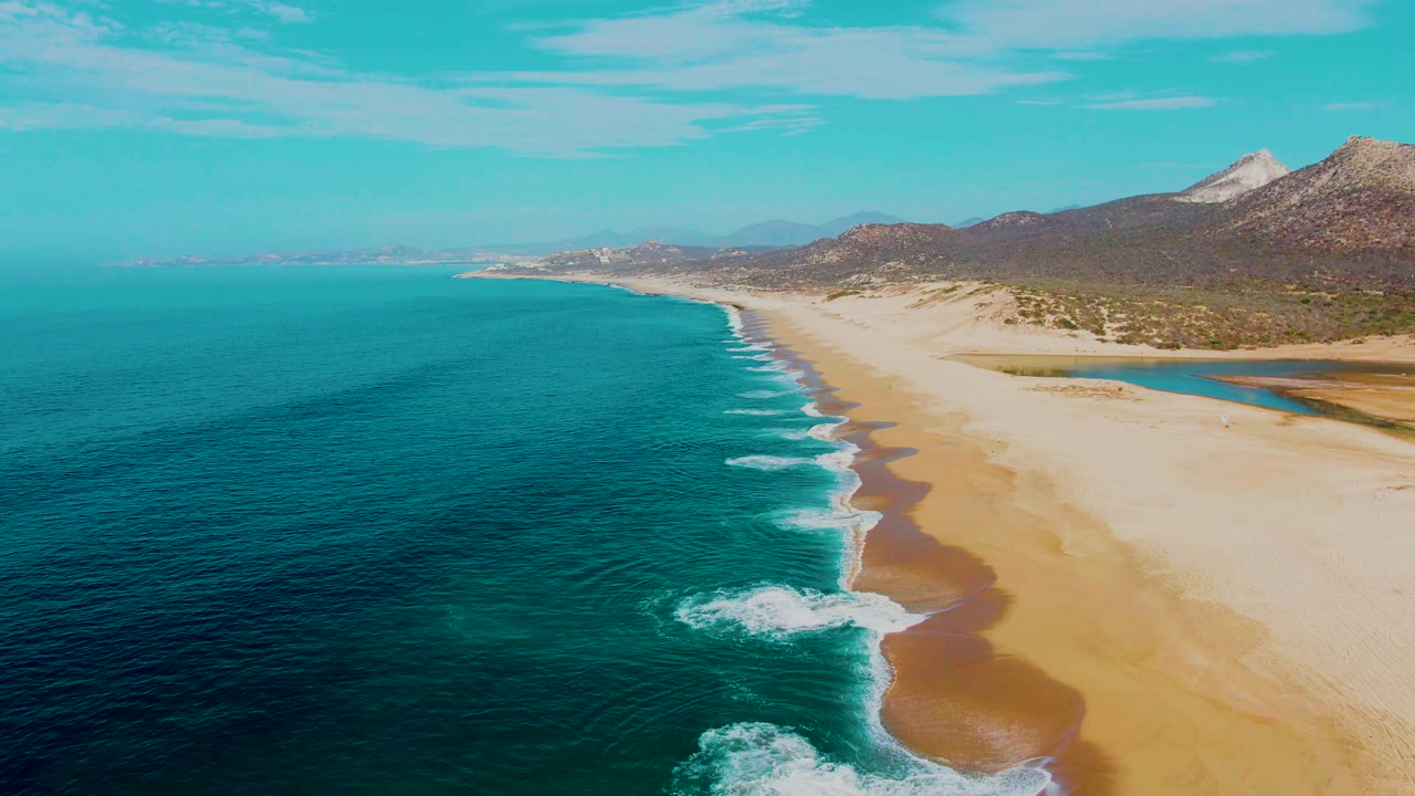 Drone-height view of Buzzards Beach, Baja California Sur, showing a diagonal surf line, pale sand, turquoise water, distant coastline, mountains, and no visible people.