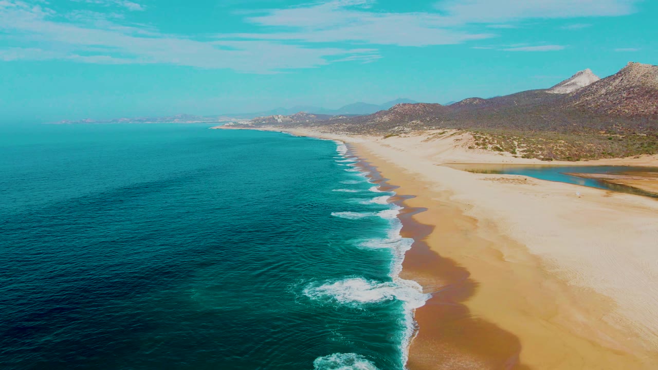 Aerial coastal frame from Punta Buzzards with white foam bands moving toward the sand, deep turquoise water offshore, dry coastal hills behind the beach, and no visible people.