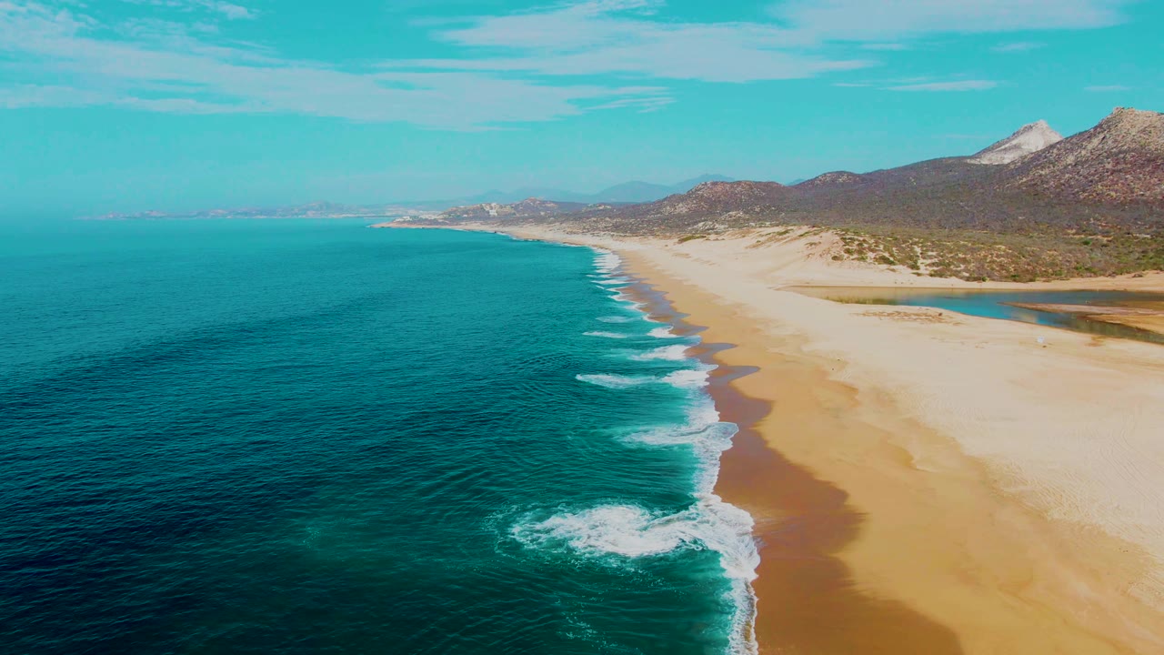Wide aerial frame over Buzzards Beach in Mexico, with foamy surf curling near the lower edge, open turquoise water to the left, sand to the right, mountains beyond, and no visible people.