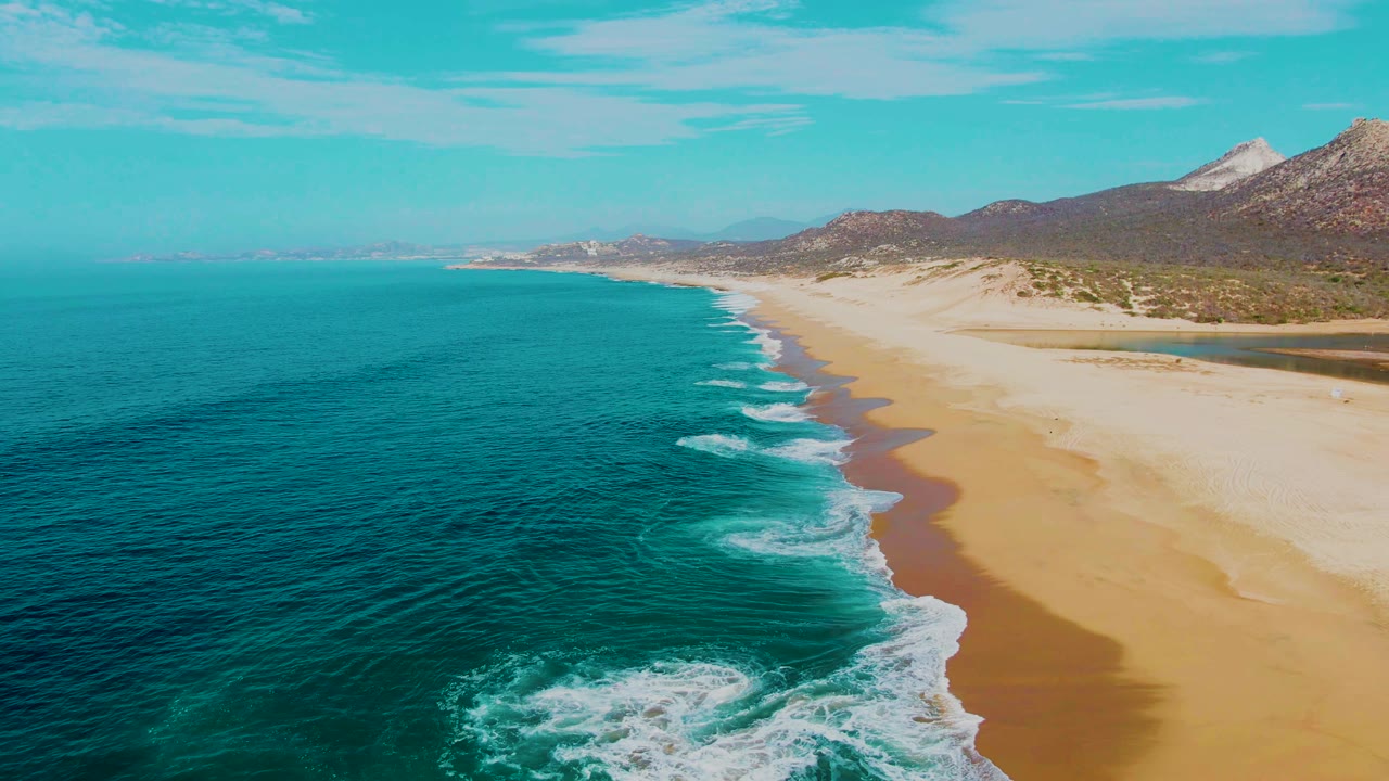 At Punta Buzzards in Baja California Sur, turquoise ocean water and white surf meet a wide sandy beach with low mountains in the distance and no visible people.