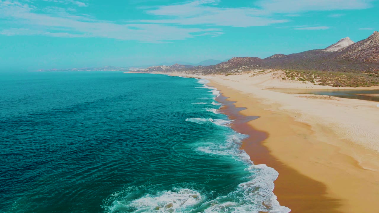 The aerial view follows the Punta Buzzards shoreline as bands of white foam curve across wet sand, with open water on the left and no visible people.