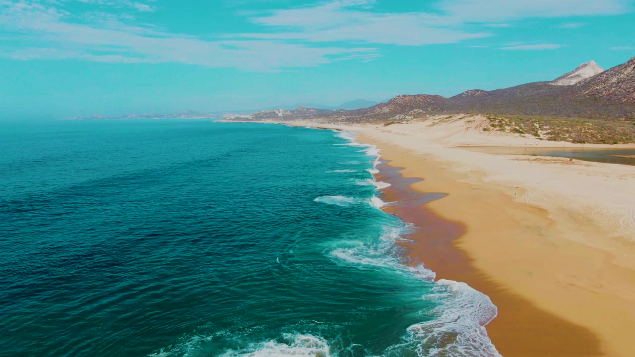 At 0:02, the surf line sits closer to the lower right edge while the open turquoise water spreads across the left side, with Baja California Sur mountains behind the beach and no visible people.