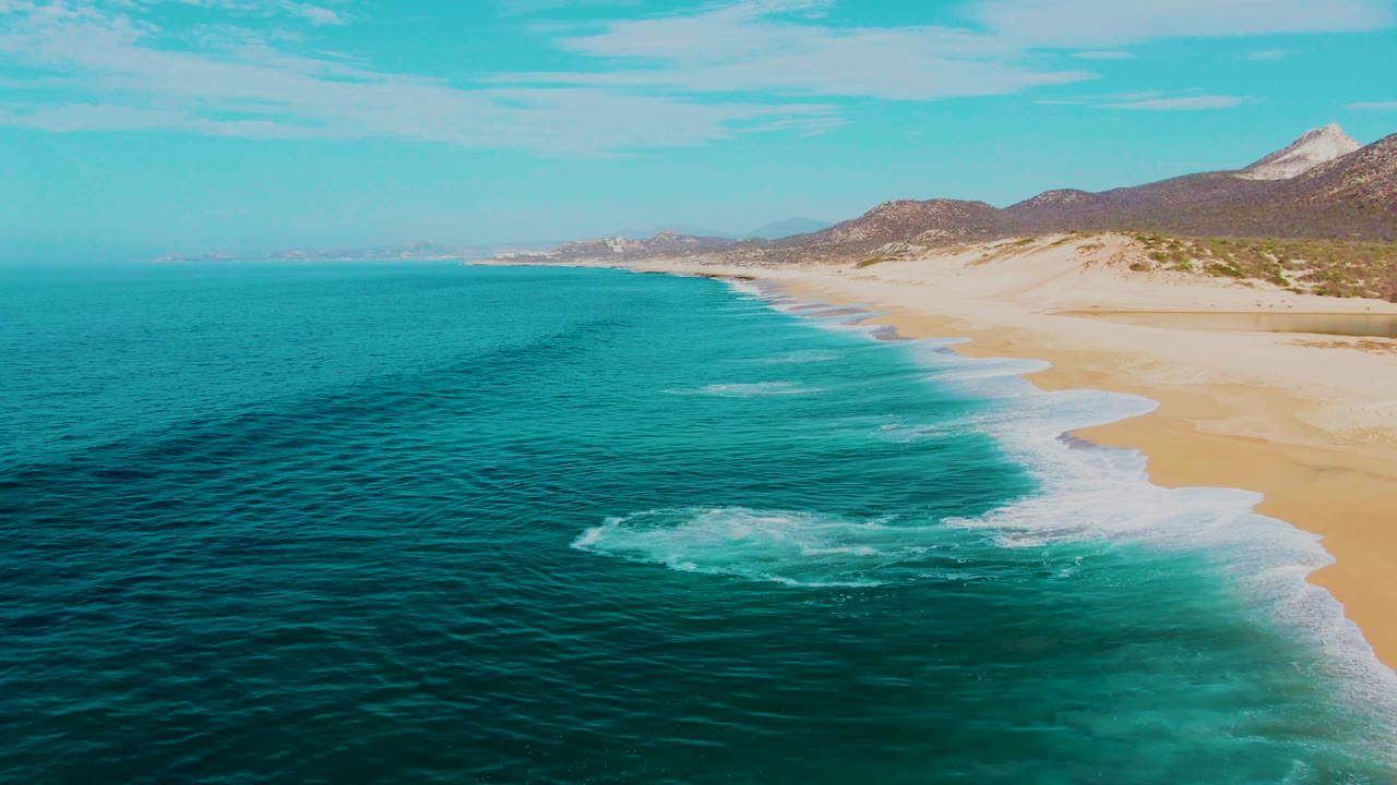 Aerial view of turquoise ocean waves reaching a sandy beach with hills beyond.