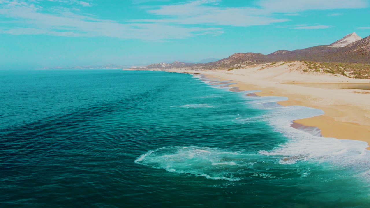 Aerial view at Buzzards Beach, East Cape, Baja California Sur, with a larger patch of white foam forming in the lower water, broad sand to the right, distant hills, and no visible people.