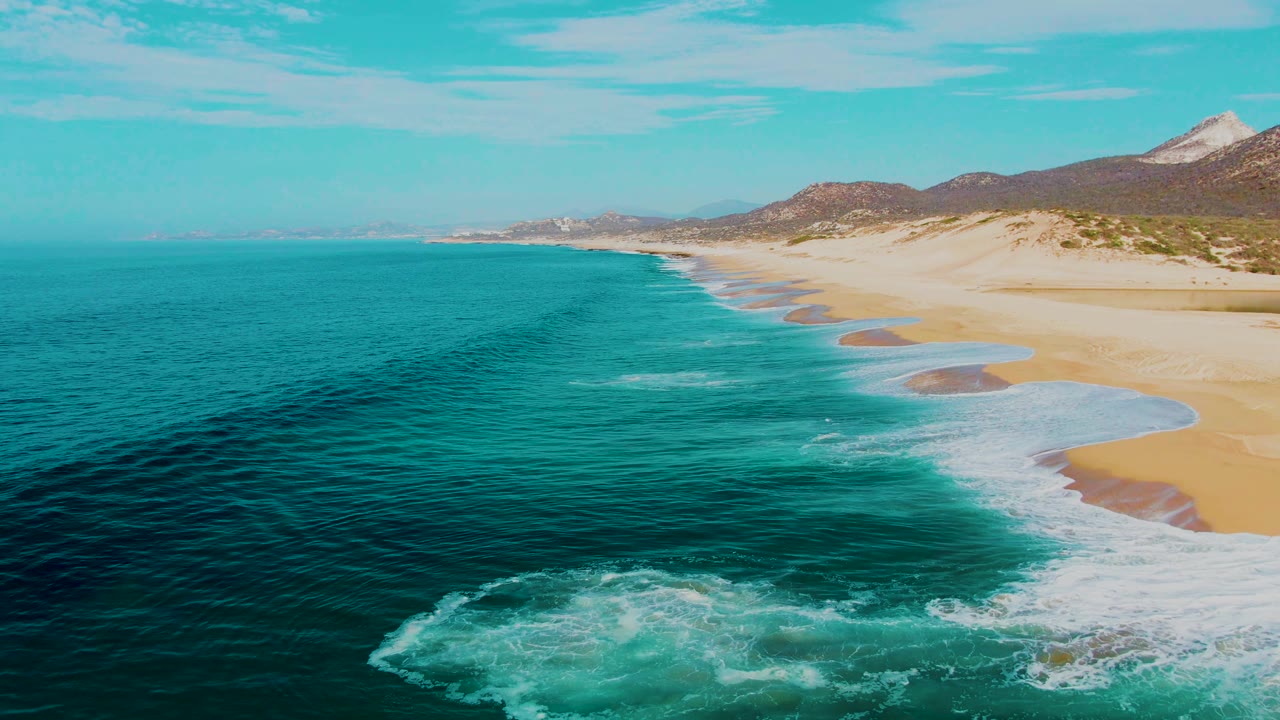 Buzzards Beach aerial frame in Baja California Sur showing the surf line closer to the lower right edge, teal water filling most of the frame, pale sand along the coast, and no visible people.