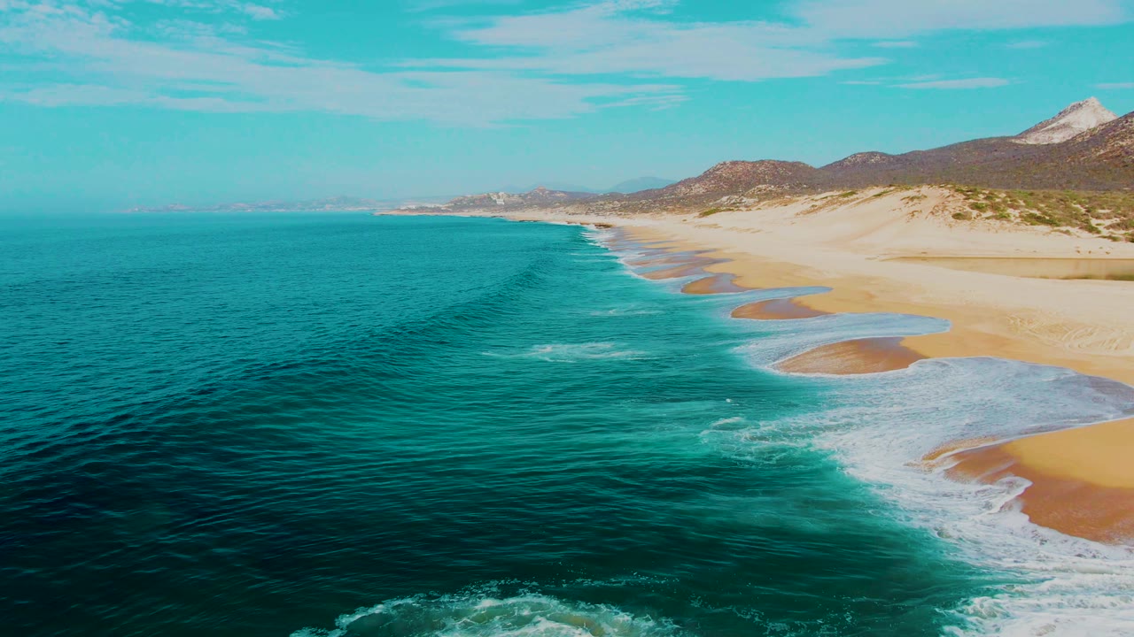 Aerial view over Buzzards Beach, East Cape, Baja California Sur, with sandbars and shallow reflective pools more prominent along the beach, open ocean to the left, and no visible people.